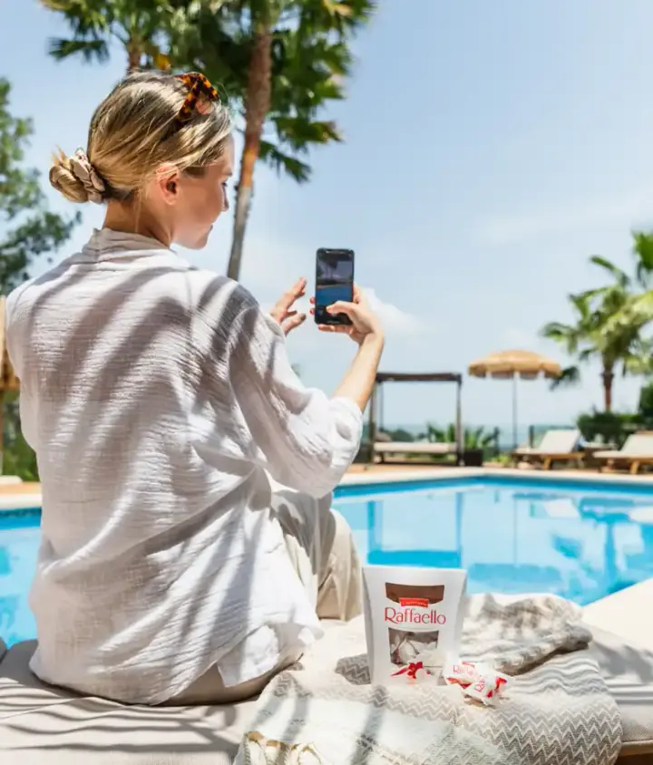 Eine Frau sitzt mit einem Smartphone am Pool; neben ihr liegt eine Süßwarenpackung auf einem Handtuch.