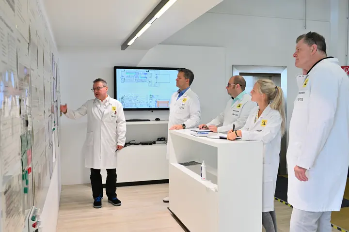 Eine Gruppe in weißen Arbeitskitteln betrachtet Pläne an einer Wand und auf einem Bildschirm.