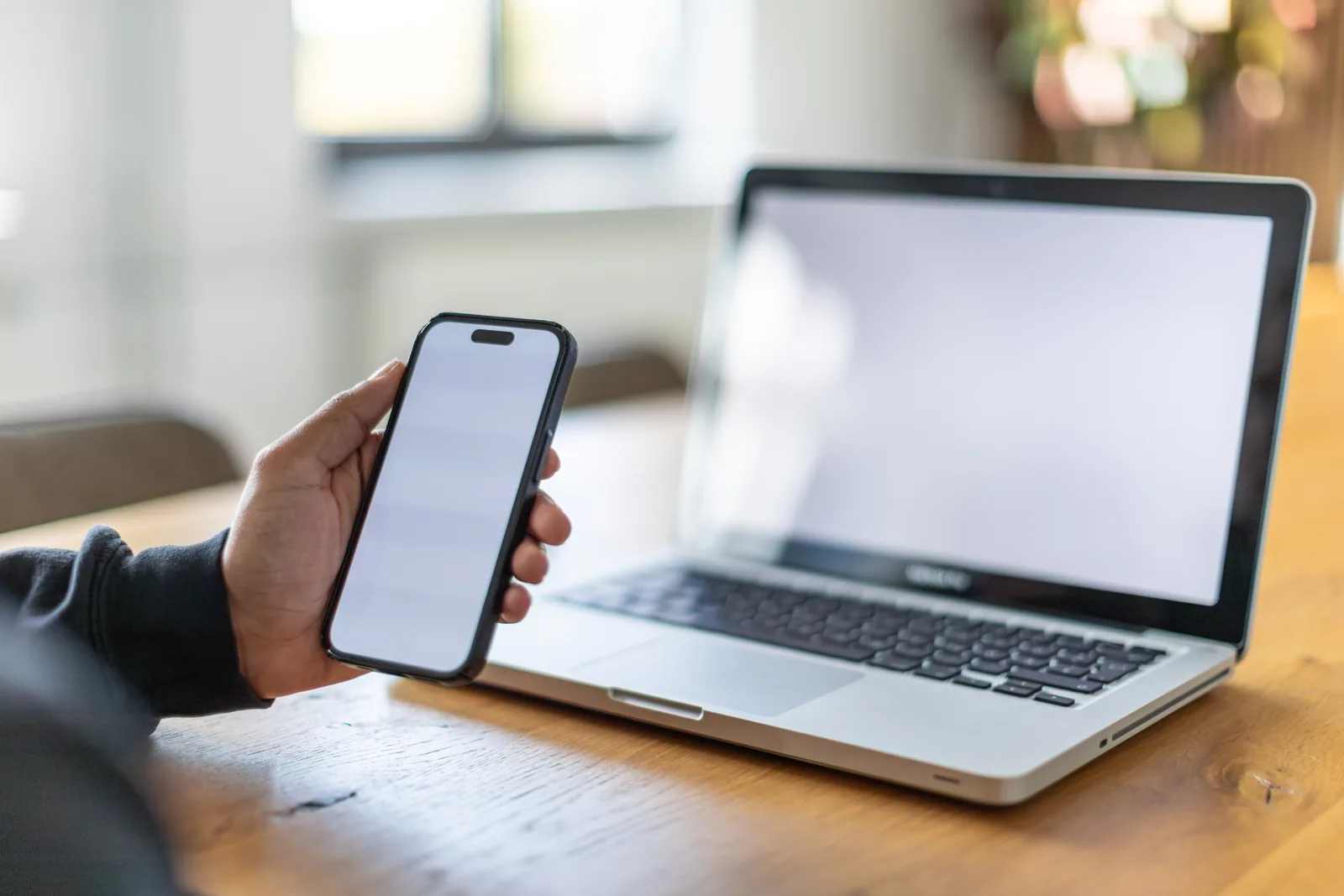 Eine Hand hält ein Smartphone vor einem geöffneten Laptop; beide Bildschirme sind ohne Inhalte.